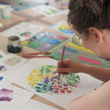 Load image into Gallery viewer, Person printing a colourful spring floral design on paper with various art supplies around.
